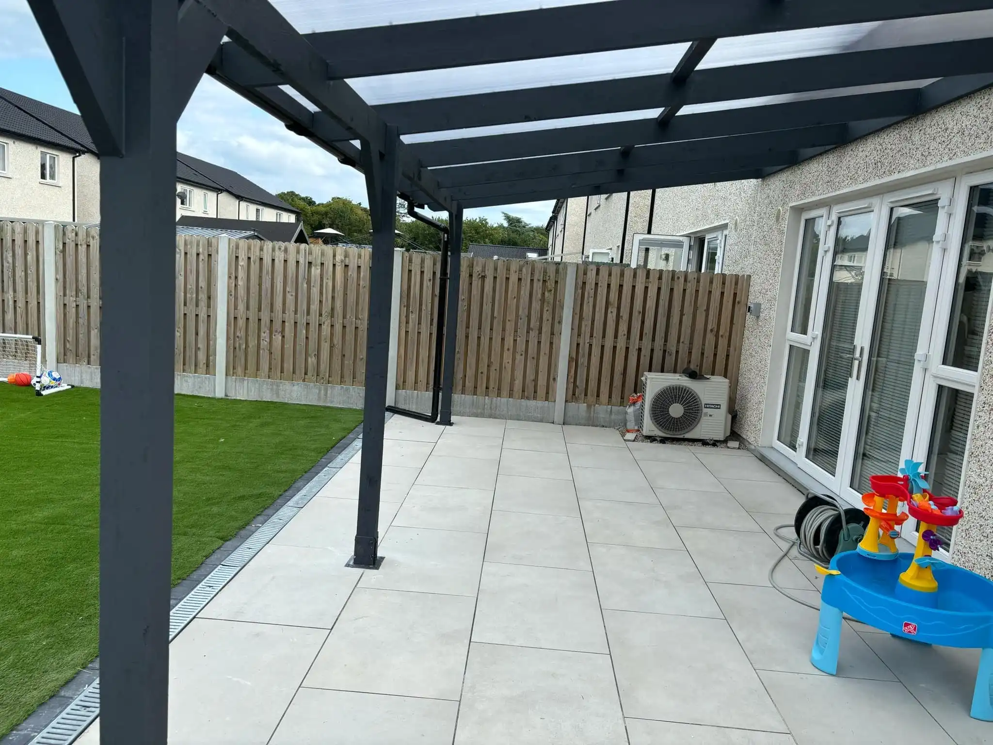 A modern lean-to pergola attached to the rear of a white two-story house, featuring a dark grey frame and a translucent polycarbonate roof. The pergola extends over a set of white double patio doors, providing shade and shelter to an outdoor living area. To the left, a dark grey shed or storage unit is integrated beneath the pergola's extension, offering practical storage. The image captures a sunny day, with a clear sky, and shows a patch of grass in the foreground. This custom pergola enhances outdoor enjoyment and functionality, blending seamlessly with the home's exterior.