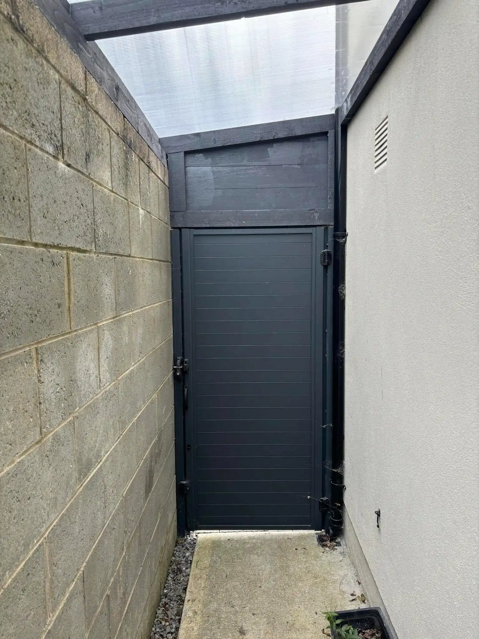 A custom-built, dark grey lean-to shed with an integrated, horizontally paneled door, perfectly fitted into a narrow side passage. The structure features a translucent lean-to roof supported by dark beams, providing shelter. This design offers secure, discreet outdoor storage and a functional, protected side entrance.