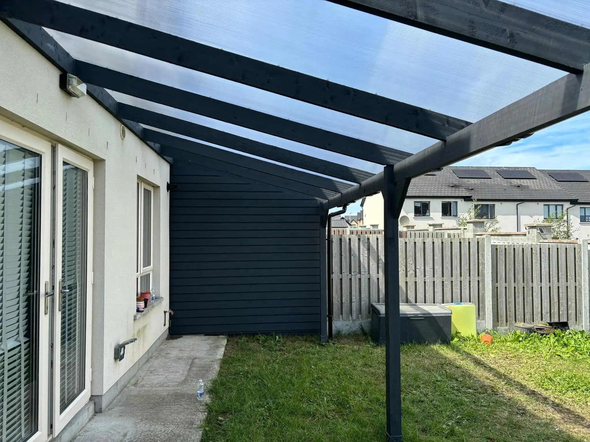 A modern lean-to pergola attached to the rear of a white two-story house, featuring a dark grey frame and a translucent polycarbonate roof. The pergola extends over a set of white double patio doors, providing shade and shelter to an outdoor living area. To the left, a dark grey shed or storage unit is integrated beneath the pergola's extension, offering practical storage. The image captures a sunny day, with a clear sky, and shows a patch of grass in the foreground. This custom pergola enhances outdoor enjoyment and functionality, blending seamlessly with the home's exterior.