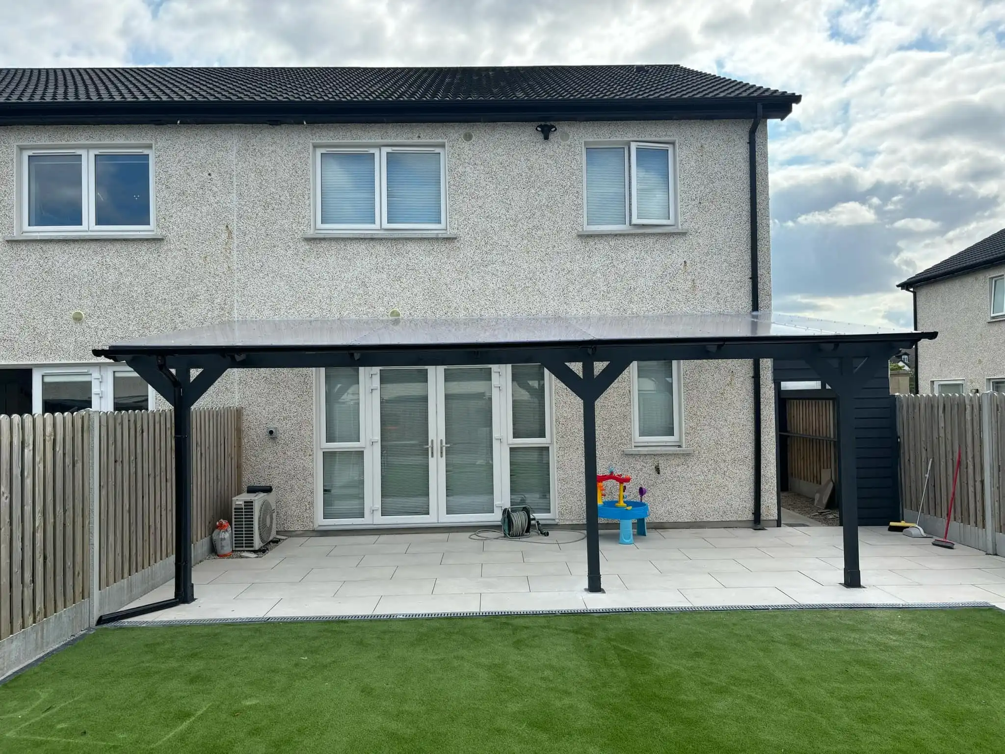 A modern lean-to pergola attached to the rear of a white two-story house, featuring a dark grey frame and a translucent polycarbonate roof. The pergola extends over a set of white double patio doors, providing shade and shelter to an outdoor living area. To the left, a dark grey shed or storage unit is integrated beneath the pergola's extension, offering practical storage. The image captures a sunny day, with a clear sky, and shows a patch of grass in the foreground. This custom pergola enhances outdoor enjoyment and functionality, blending seamlessly with the home's exterior.