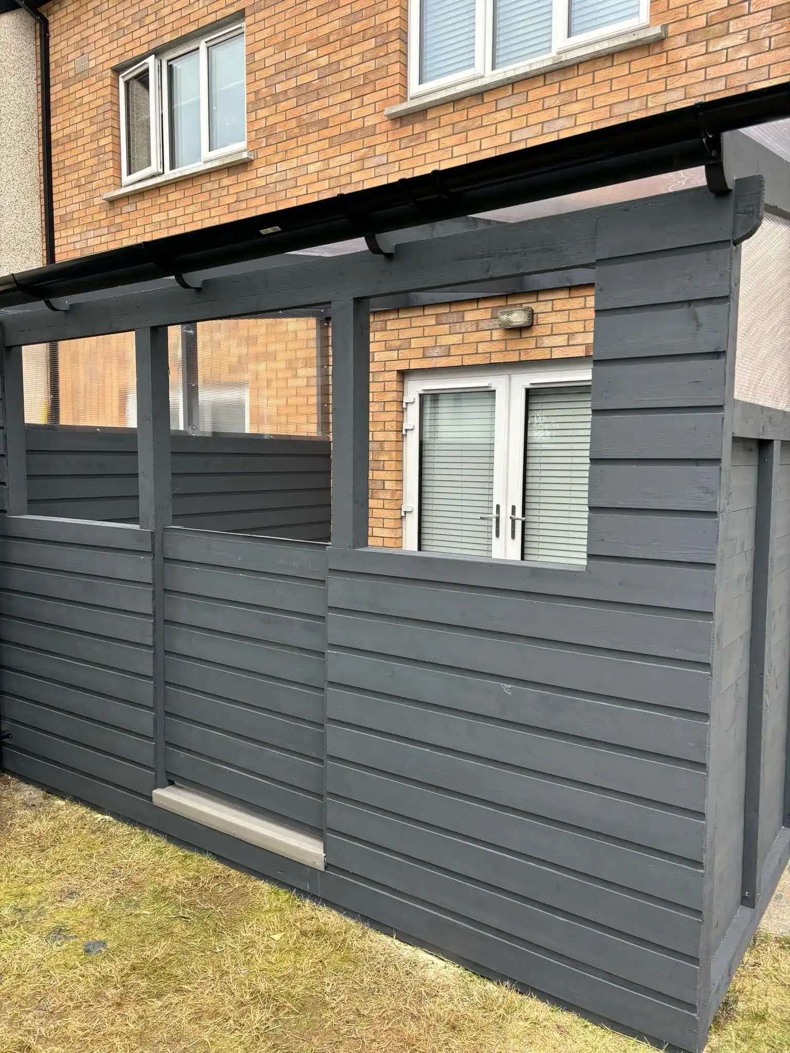 A modern dark grey horizontal slat garden partition, featuring clear transparent panels in its upper section for light while maintaining privacy. The structure is integrated with a black gutter system along the top, suggesting it's part of a lean-to shelter or pergola system. The background shows the brick exterior of a house with white patio doors and windows, and a patch of green grass is visible in the foreground. This garden feature provides both privacy and an element of design, suitable for defining outdoor spaces.