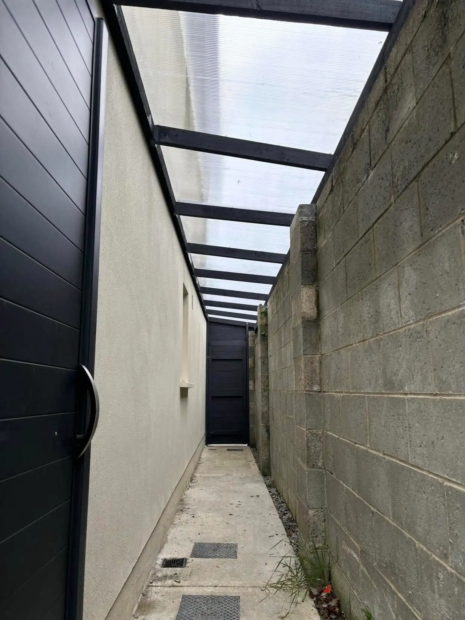 A custom-built, dark grey lean-to shed with an integrated, horizontally paneled door, perfectly fitted into a narrow side passage. The structure features a translucent lean-to roof supported by dark beams, providing shelter. This design offers secure, discreet outdoor storage and a functional, protected side entrance.