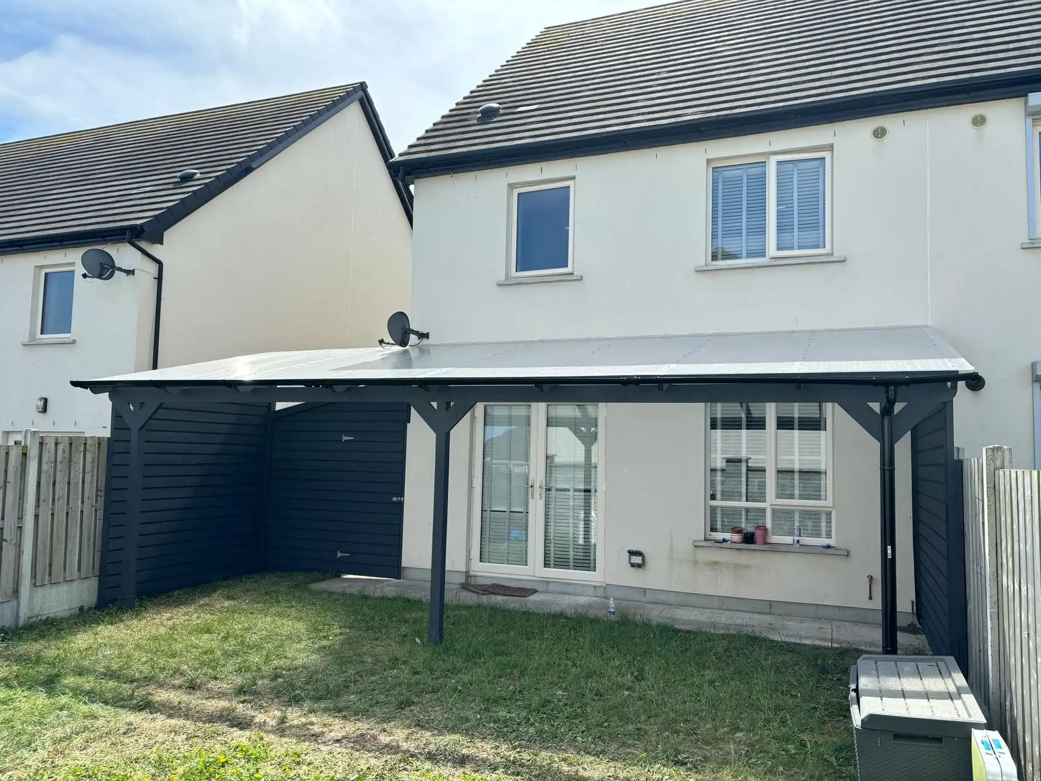 A modern lean-to pergola attached to the rear of a white two-story house, featuring a dark grey frame and a translucent polycarbonate roof. The pergola extends over a set of white double patio doors, providing shade and shelter to an outdoor living area. To the left, a dark grey shed or storage unit is integrated beneath the pergola's extension, offering practical storage. The image captures a sunny day, with a clear sky, and shows a patch of grass in the foreground. This custom pergola enhances outdoor enjoyment and functionality, blending seamlessly with the home's exterior.