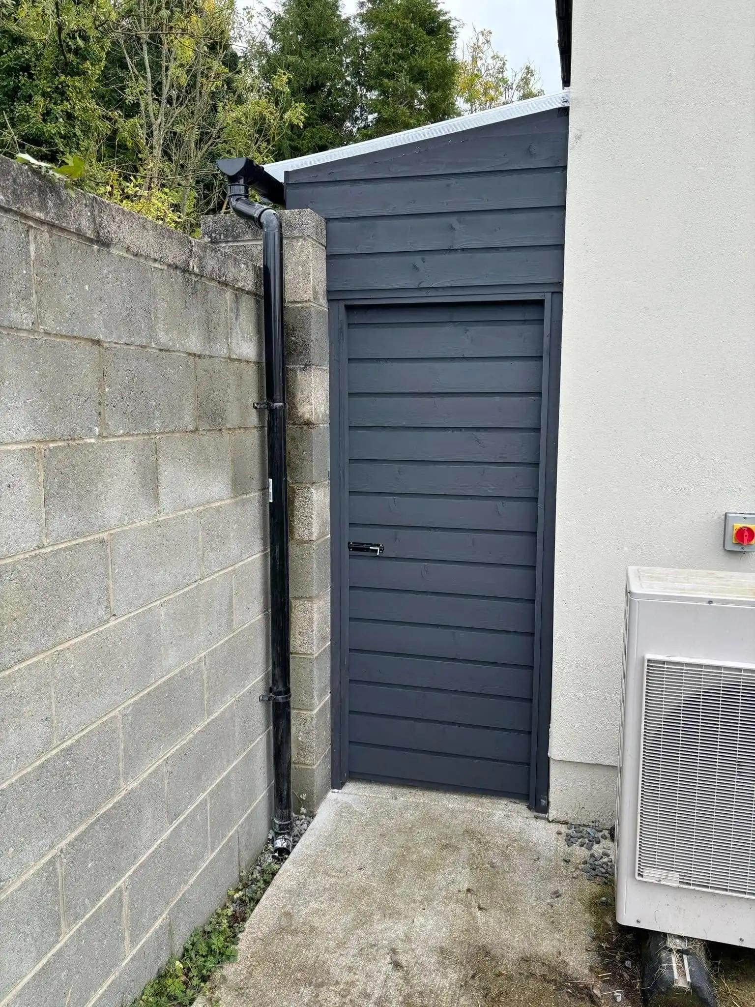 A custom-built, dark grey lean-to shed with an integrated, horizontally paneled door, perfectly fitted into a narrow side passage. The structure features a translucent lean-to roof supported by dark beams, providing shelter. This design offers secure, discreet outdoor storage and a functional, protected side entrance.